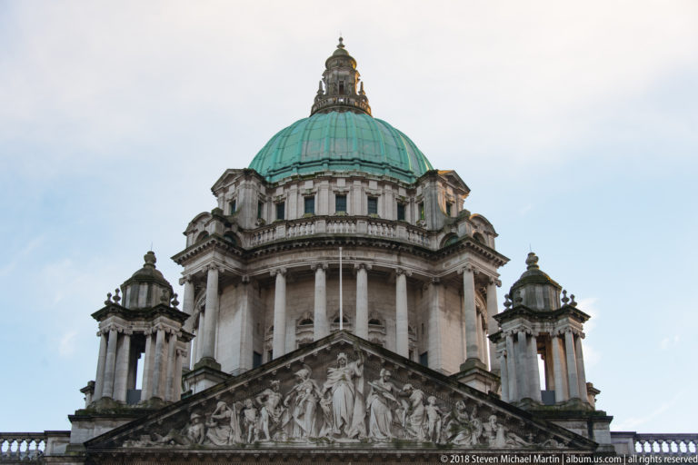 Low-angle view of Belfast City Hall’s sculpted pediment, columned dome drum, green copper dome, and smaller corner pavilions.