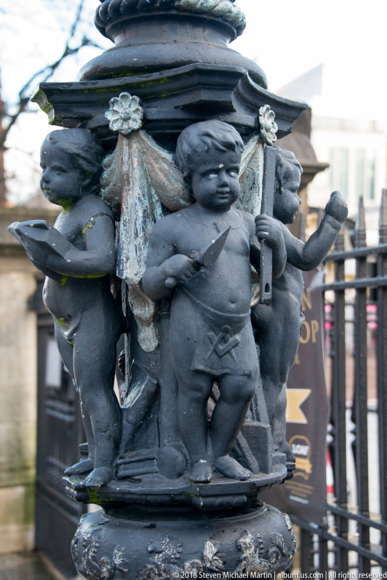 Cast-metal cherubs with industrial attributes encircling the weathered base of an ornamental lamp standard at Belfast City Hall.