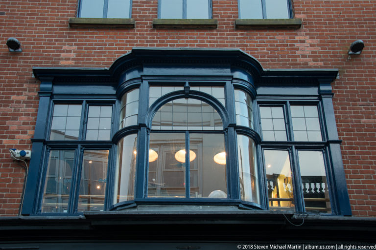 Dark-painted projecting bay window with arched central glazing and warm interior lights set into a red-brick wall.
