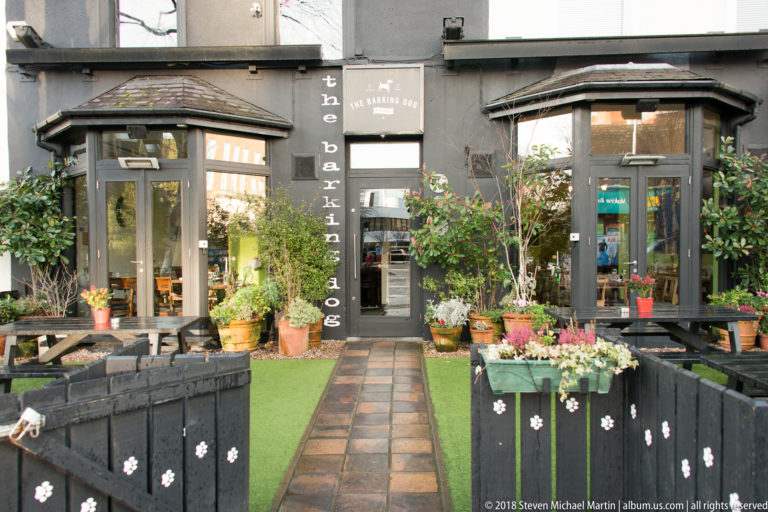 Entrance to The Barking Dog pub, framed by dark glazed bays, potted plants, flowers, and a narrow green walkway.