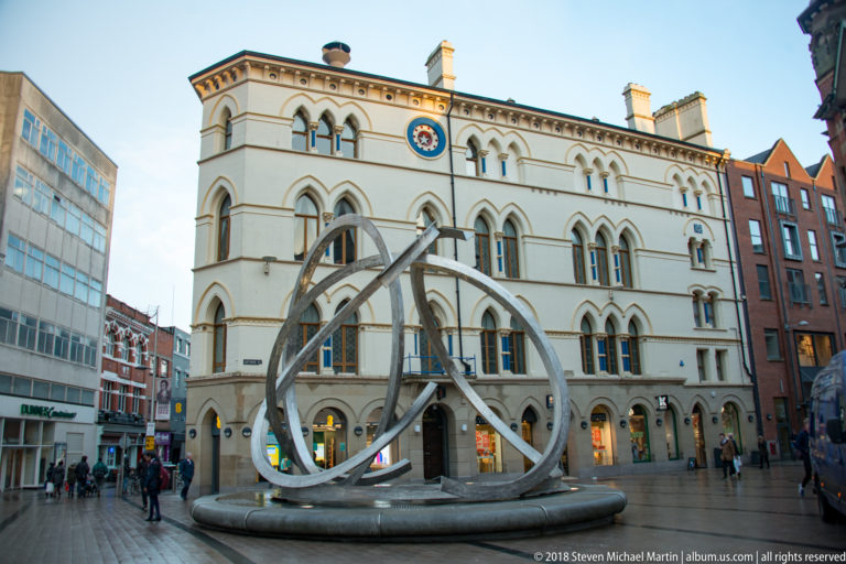 Spirit of Belfast stainless-steel sculpture forming large intertwined loops in a city-centre square before a historic cream facade.