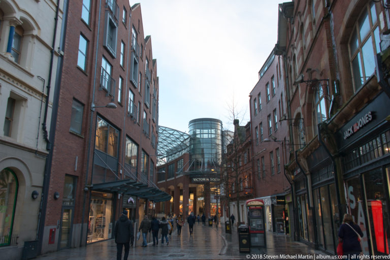 Pedestrians walk between brick shopfronts toward the glass-covered Victoria Square complex in central Belfast.
