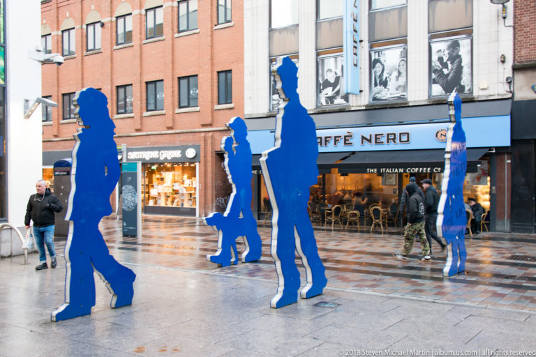 Bright-blue walking human silhouettes stand on a wet pedestrian plaza in front of Belfast shops.