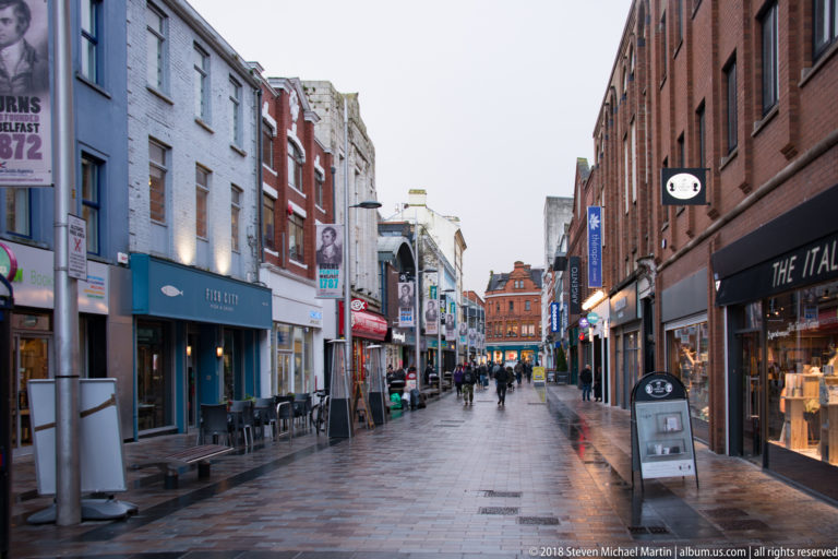 Wet pedestrian shopping lane lined with restaurants and shops in central Belfast.