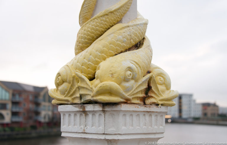 Cluster of carved fish twisting upward atop a pale architectural post near the Belfast waterfront.