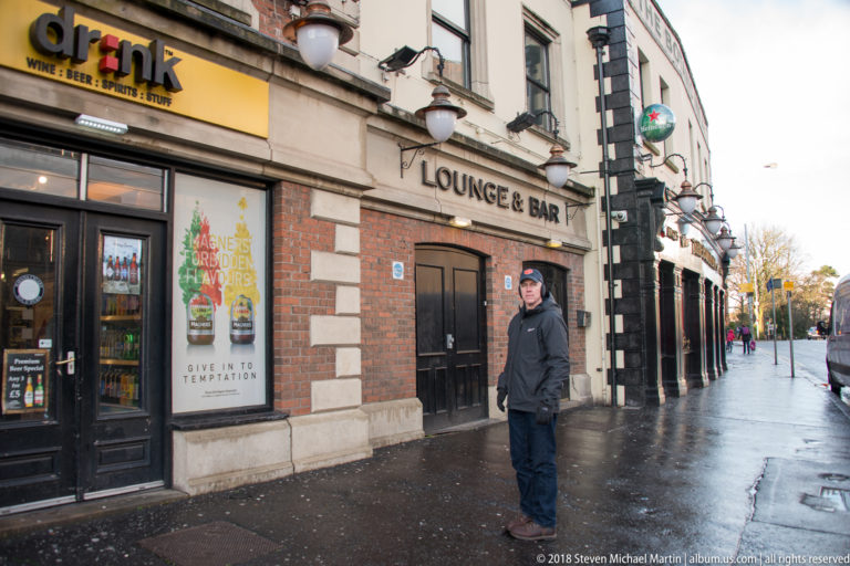 Red-brick pub exterior marked “LOUNGE & BAR,” with globe lamps, a drinks poster, and a man on wet pavement.
