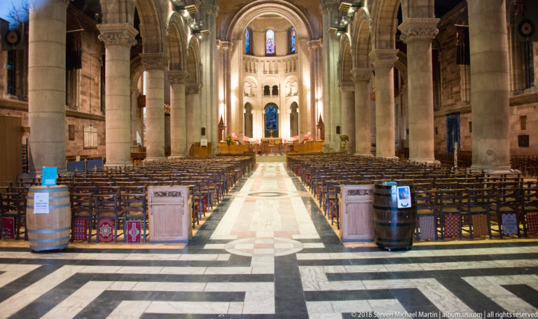 Central nave of St Anne's Cathedral with rows of chairs, massive columns, round arches, and stained glass.