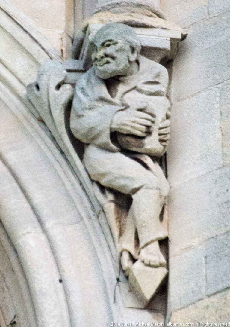 Seated weathered stone figure holding an object across its lap on St Anne's Cathedral.