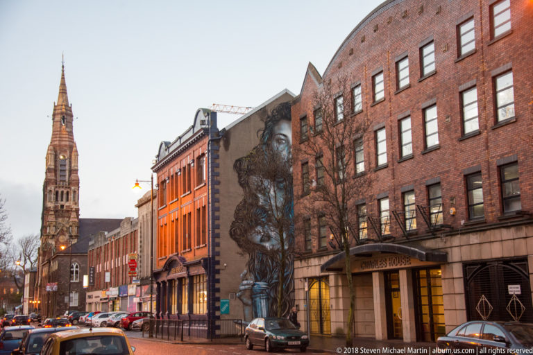 Belfast street lined with red-brick buildings, a tall church spire, and a large mural on a gable wall.