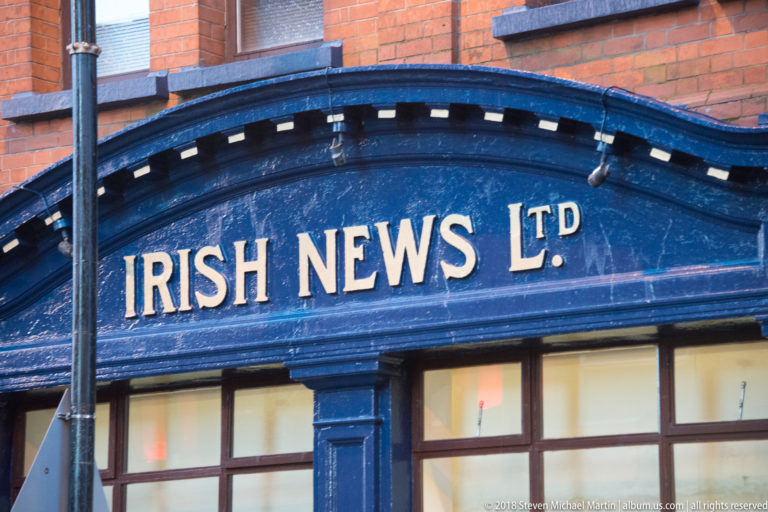 Gold IRISH NEWS LTD lettering on a dark blue painted storefront fascia beneath red brickwork.