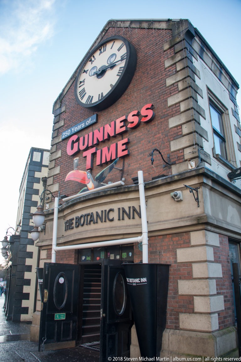 Low-angle exterior of The Botanic Inn with a large clock, “GUINNESS TIME” sign, and dark pub entrance below.