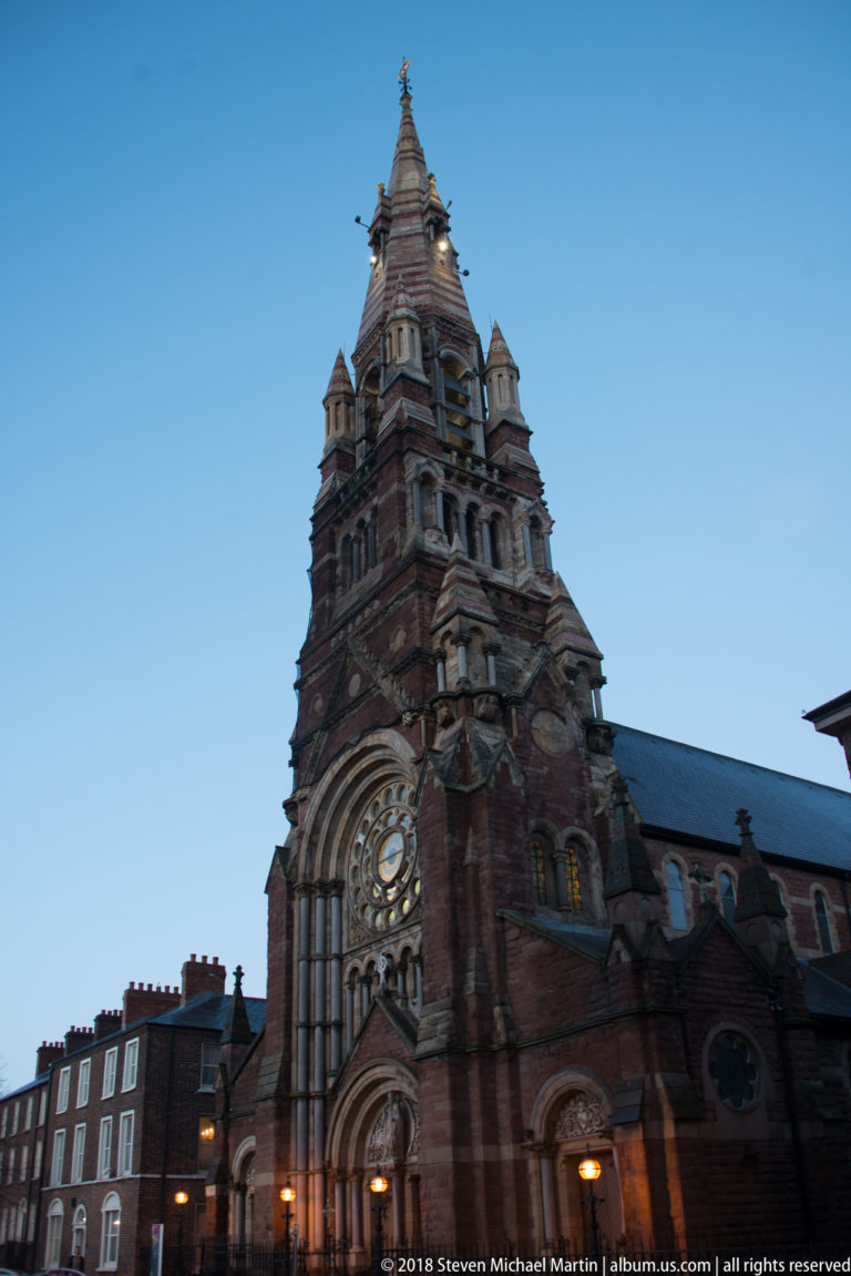 St Patrick’s Church on Donegall Street at dusk, with red-sandstone west front, rose window, tower, and 180-foot spire.