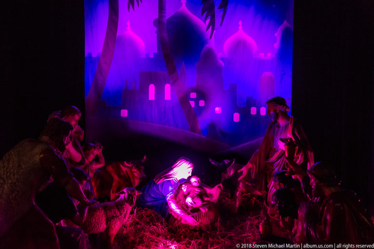 Illuminated Nativity scene with Mary, infant Jesus, Joseph, and surrounding figures under purple and blue lighting.