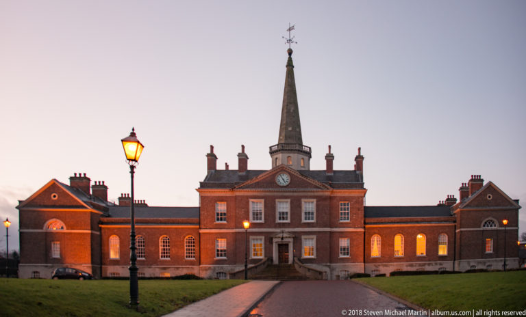 Symmetrical red-brick Georgian building with central entrance, lit windows, lamppost, and a slender spire behind.