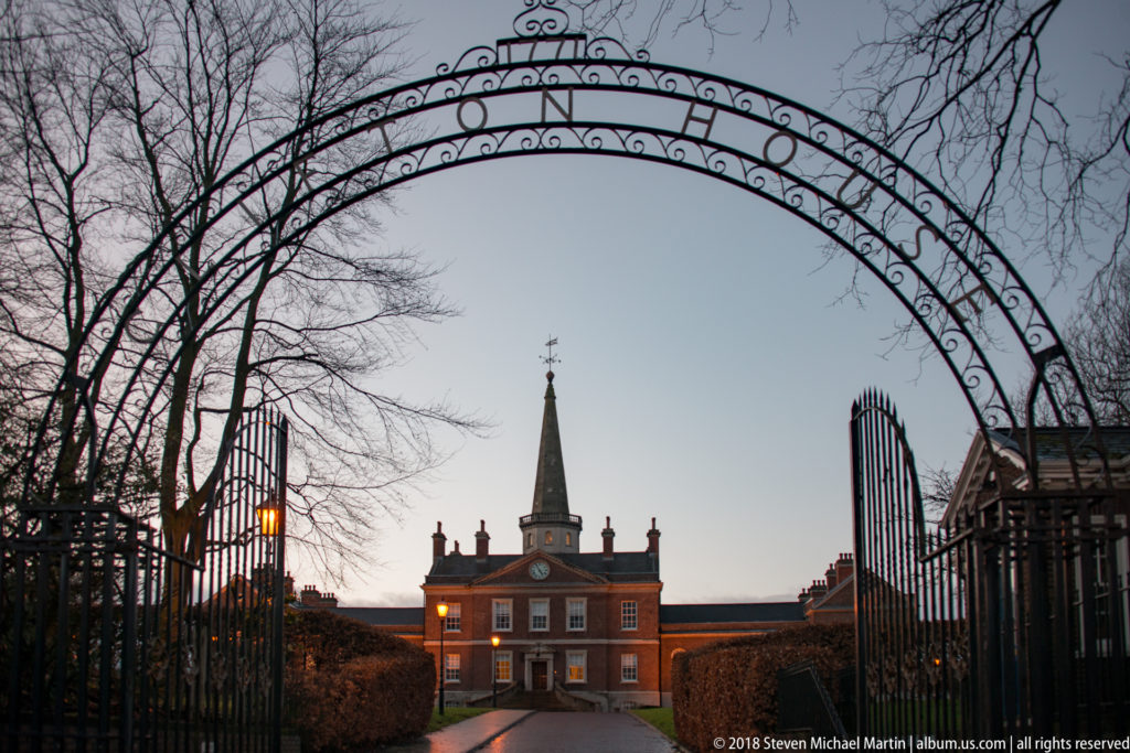 Decorative wrought-iron gateway frames a symmetrical red-brick Georgian building at twilight.