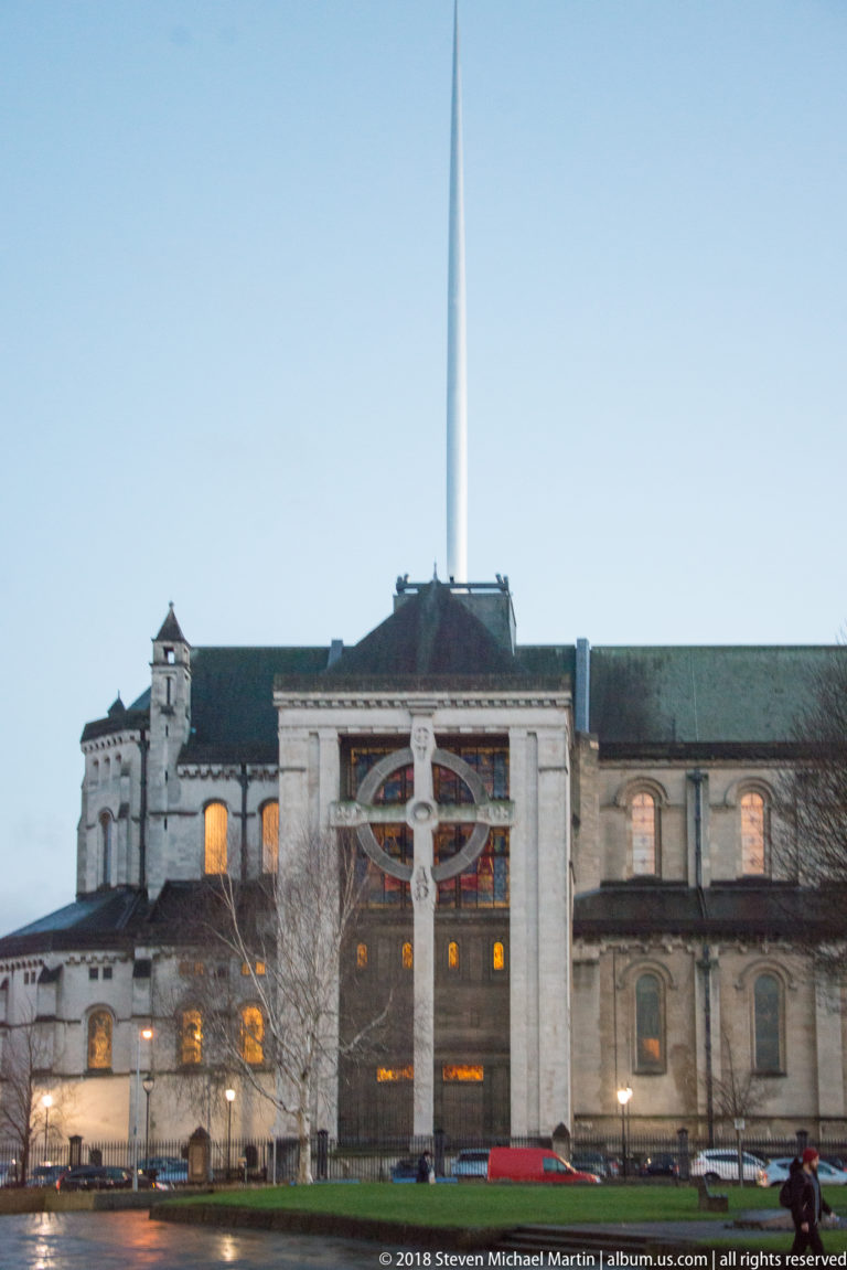 Large stone Belfast building behind a Celtic cross, with a very slender spire rising above the roofline.
