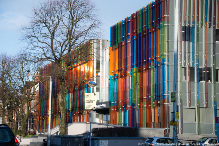 Modern building wrapped in closely spaced vertical fins in red, blue, orange, green, and gray, seen beside trees.