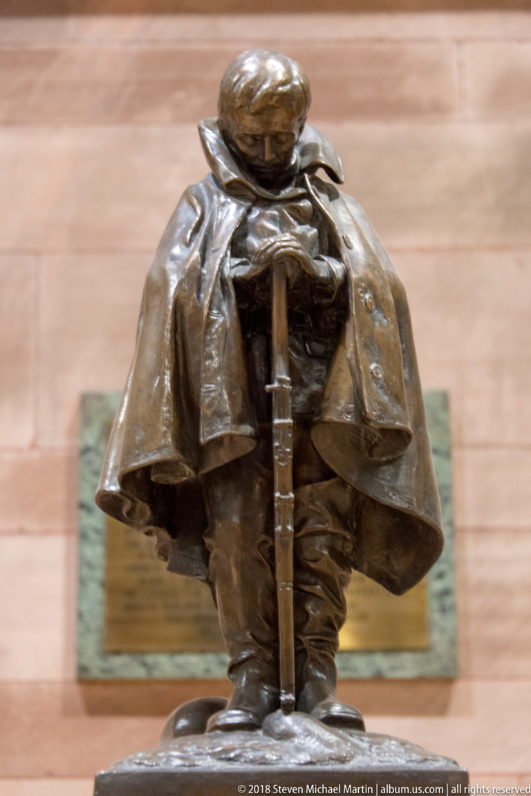 Bronze sculpture of a cloaked elderly figure seated with both hands resting on a walking stick.