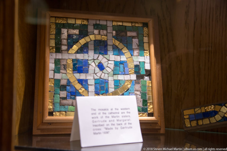 Framed blue, green, white, and gold mosaic panel beside an interpretation card in a wooden display case.