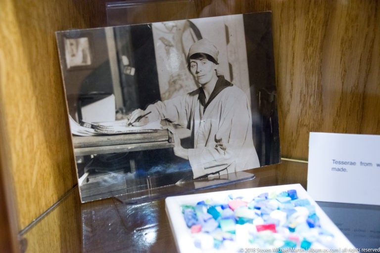 Archival photograph of a woman at a work table above a tray of colorful loose mosaic tesserae.