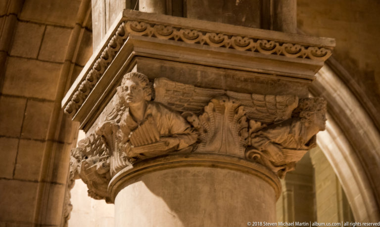 Carved stone capital with human and winged figures above a cylindrical cathedral pier.