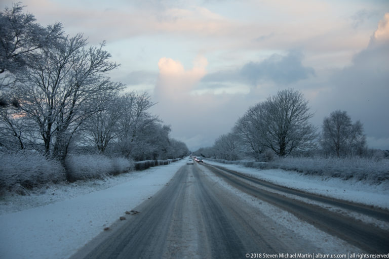 Snow-covered two-lane road lined with frosted hedges and bare trees beneath a pale winter sky.