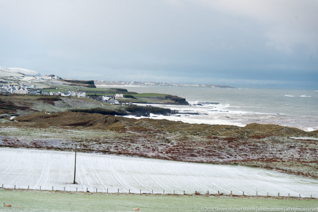 Elevated view over snowy fields, coastal buildings, rocky headlands, and breaking Atlantic surf.