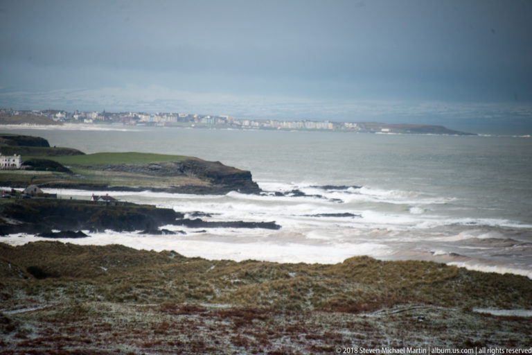 Rocky headlands and white surf in the foreground, with a distant coastal settlement across gray water.