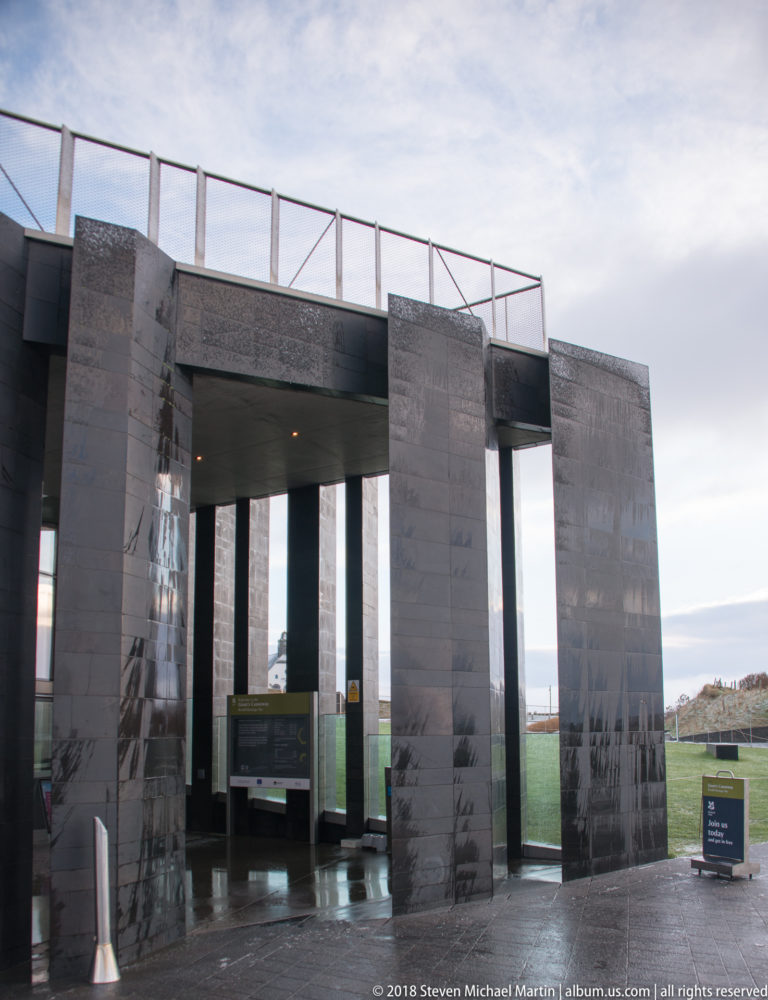 Dark stone-clad visitor-centre fins and glazed entrance opening with the coastal landscape visible beyond.