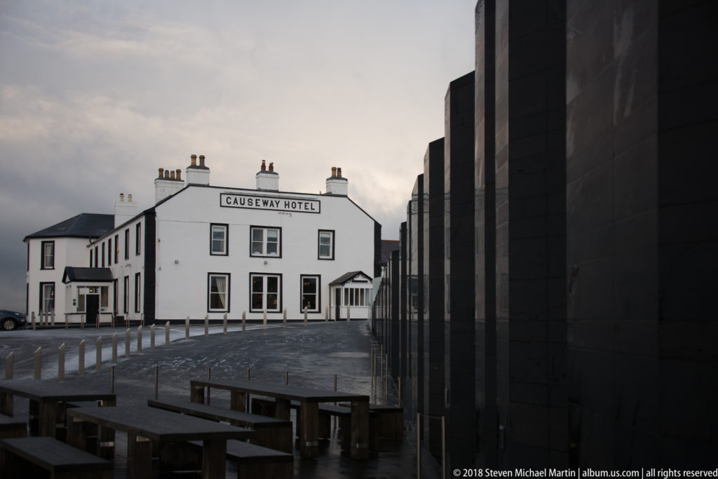 White Causeway Hotel façade seen beyond dark visitor-centre walls and outdoor benches.