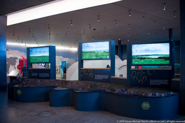 Curved visitor-centre exhibition with landscape screens, maps, interpretive panels, and geology displays beneath ceiling spotlights.