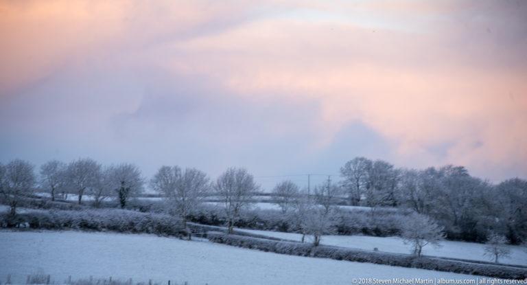 Snow-covered rolling fields divided by hedges and leafless trees beneath soft pink and blue clouds.