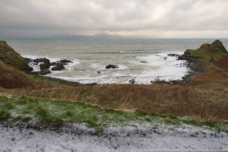 Rocky cove between grassy slopes with white Atlantic surf and patches of snow in the foreground.