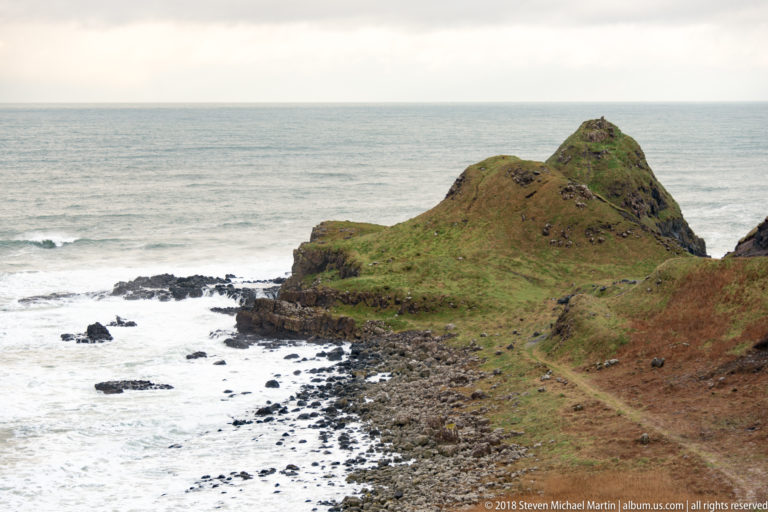 Steep grassy headland above a rocky shore and white surf, with a narrow footpath crossing the slope.