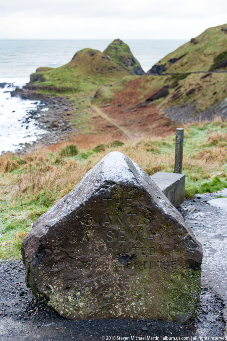 Wet, weathered stone marker beside the path with partially legible Giant’s Causeway lettering and grassy slopes behind.