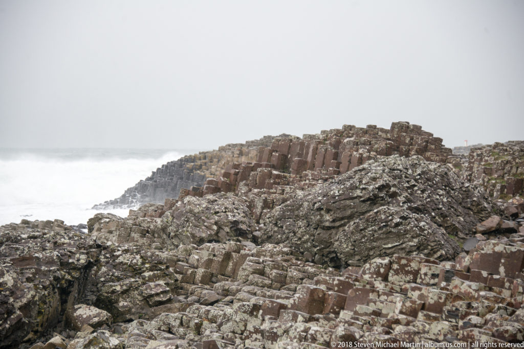 Low ridge of columnar basalt behind fractured volcanic rocks under a pale gray sky.