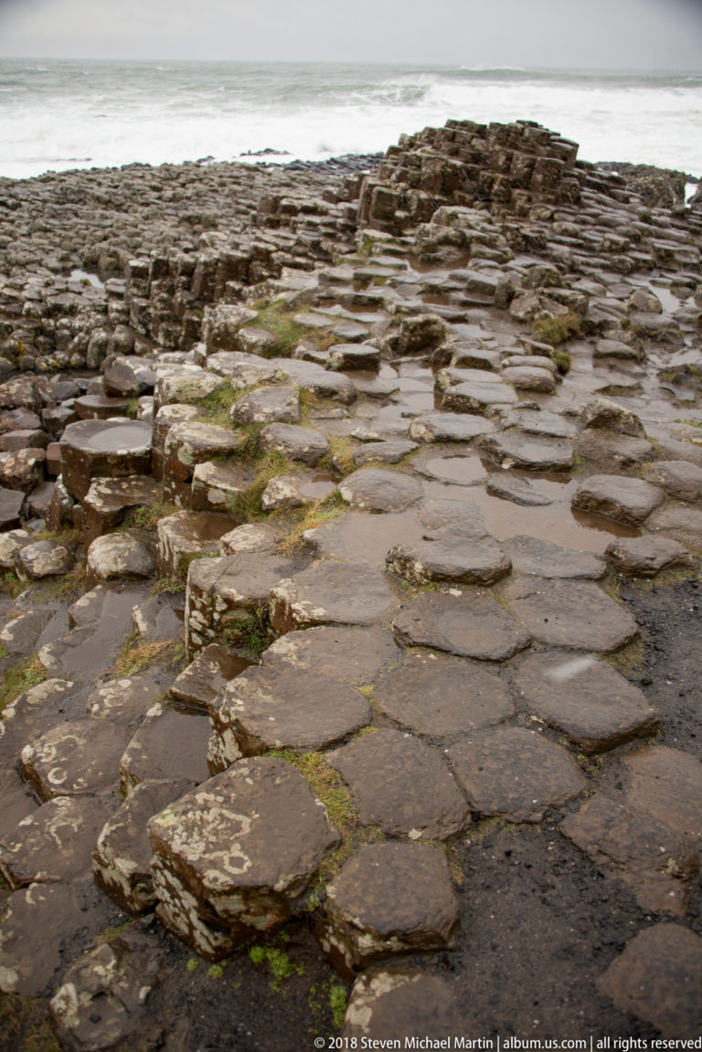 Wet polygonal basalt column tops forming a natural pavement with shallow puddles and the sea beyond.