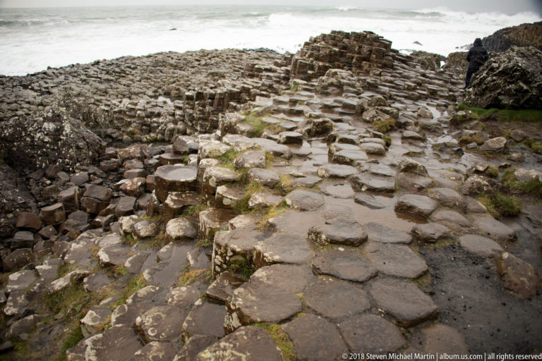 Uneven wet basalt columns with polygonal tops rising in steps toward a higher cluster near the sea.