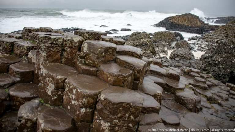 Large wet polygonal basalt columns in the foreground, descending toward lower rocks and rough Atlantic surf.