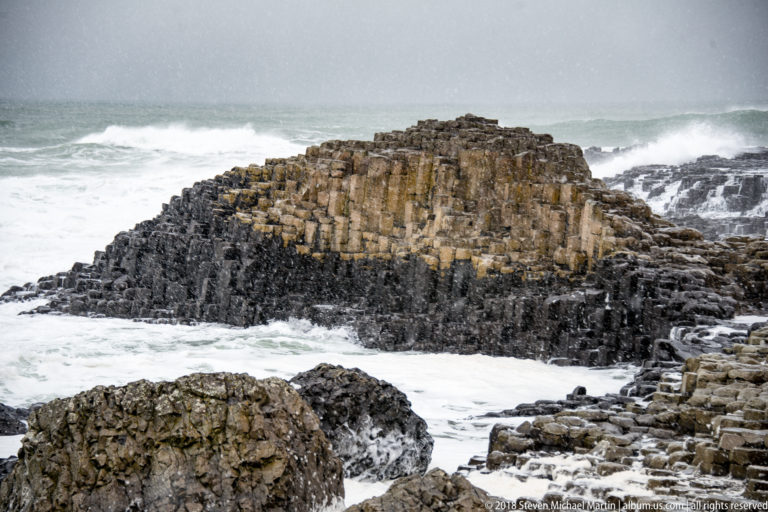 Low columnar-basalt outcrop surrounded by white surf, with dark rocks in front and wintry spray or precipitation.