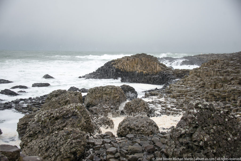 Basalt boulders and columnar ledges along the shore, with a larger outcrop beyond in white Atlantic surf.