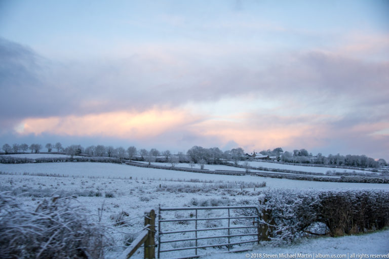Metal farm gate before snow-covered fields, hedgerows, fences, and distant houses under a pastel winter sky.