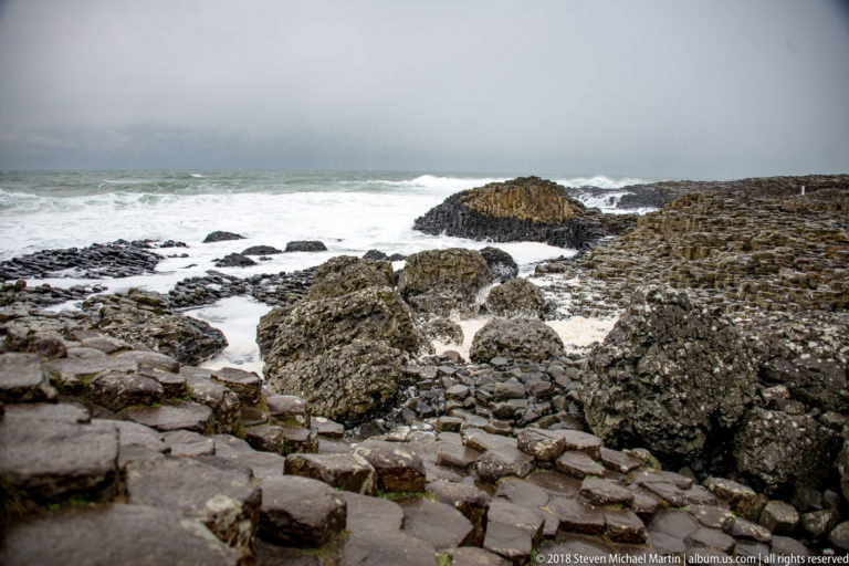 Rocky basalt shelves and blocks surrounding white surf, with a low columnar outcrop in the middle distance.