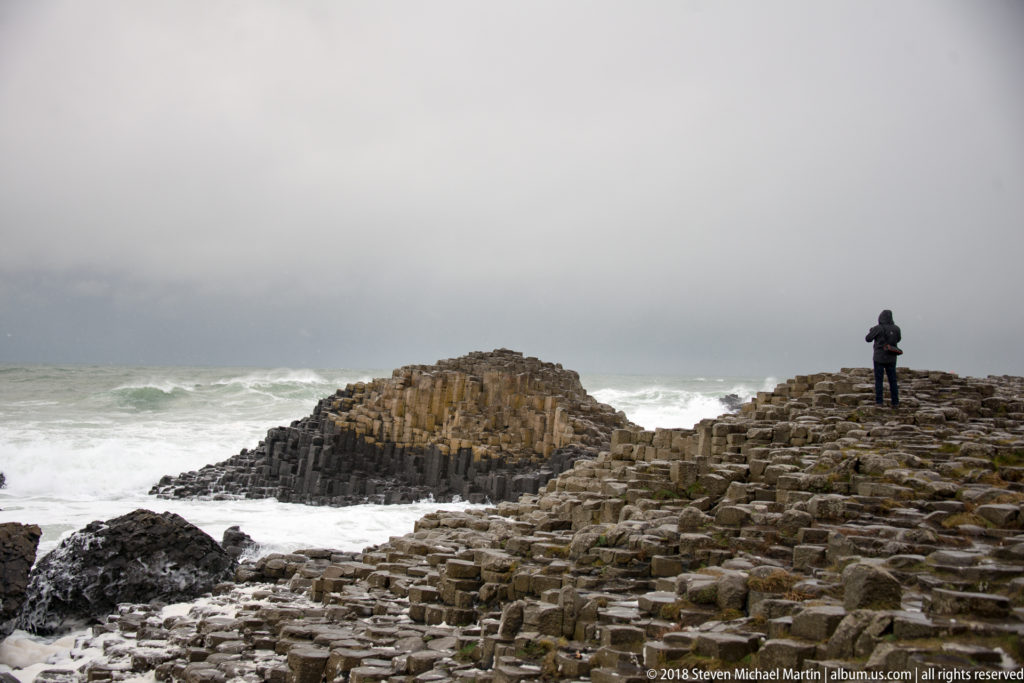 Visitor standing on wet Giant’s Causeway basalt beside a raised columnar outcrop and rough white Atlantic surf.