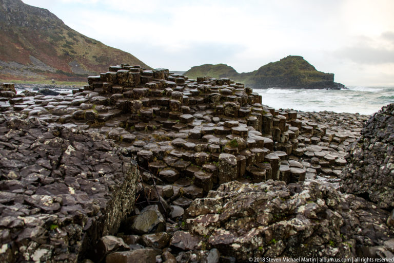 Stepped basalt mound among fractured rocks and puddles, with rough sea and a dark offshore headland beyond.