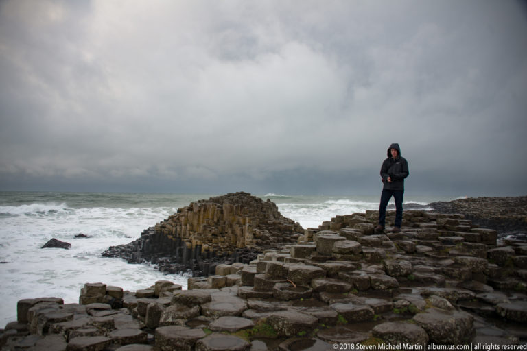 Visitor standing on Giant’s Causeway basalt beside a raised column formation with large waves behind.