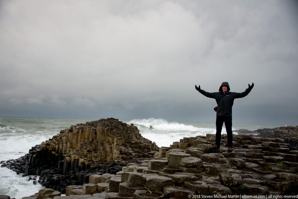 Visitor with both arms raised on wet basalt beside a compact column mound and breaking Atlantic waves.