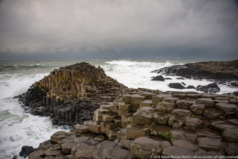 Low ochre-gray basalt mound surrounded by white surf, with wet polygonal blocks across the foreground.