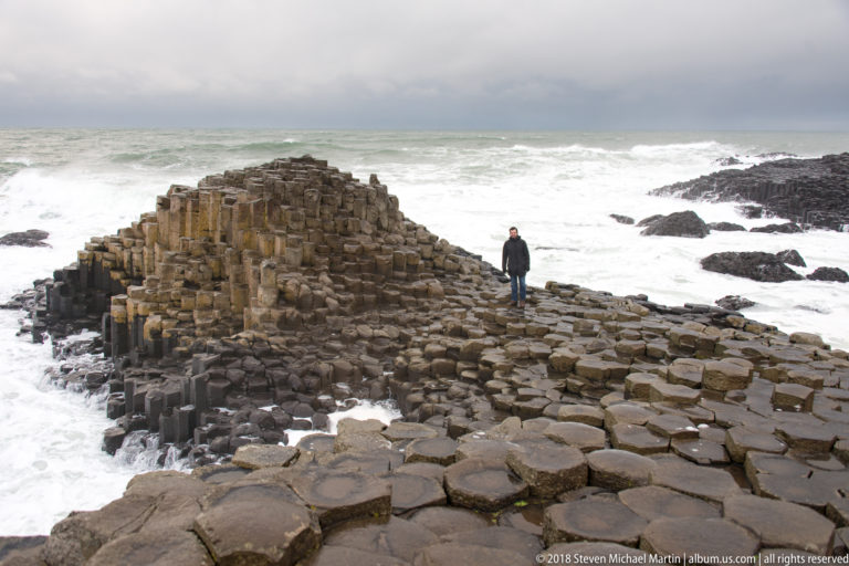 Visitor standing beside a tall basalt mound, with broad polygonal column tops and white surf surrounding the formation.