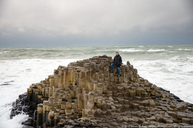 Visitor seated atop a steep basalt mound with white surf and gray-green Atlantic water behind.